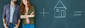 Couple standing in front of a chalkboard with chalk drawing of a house.